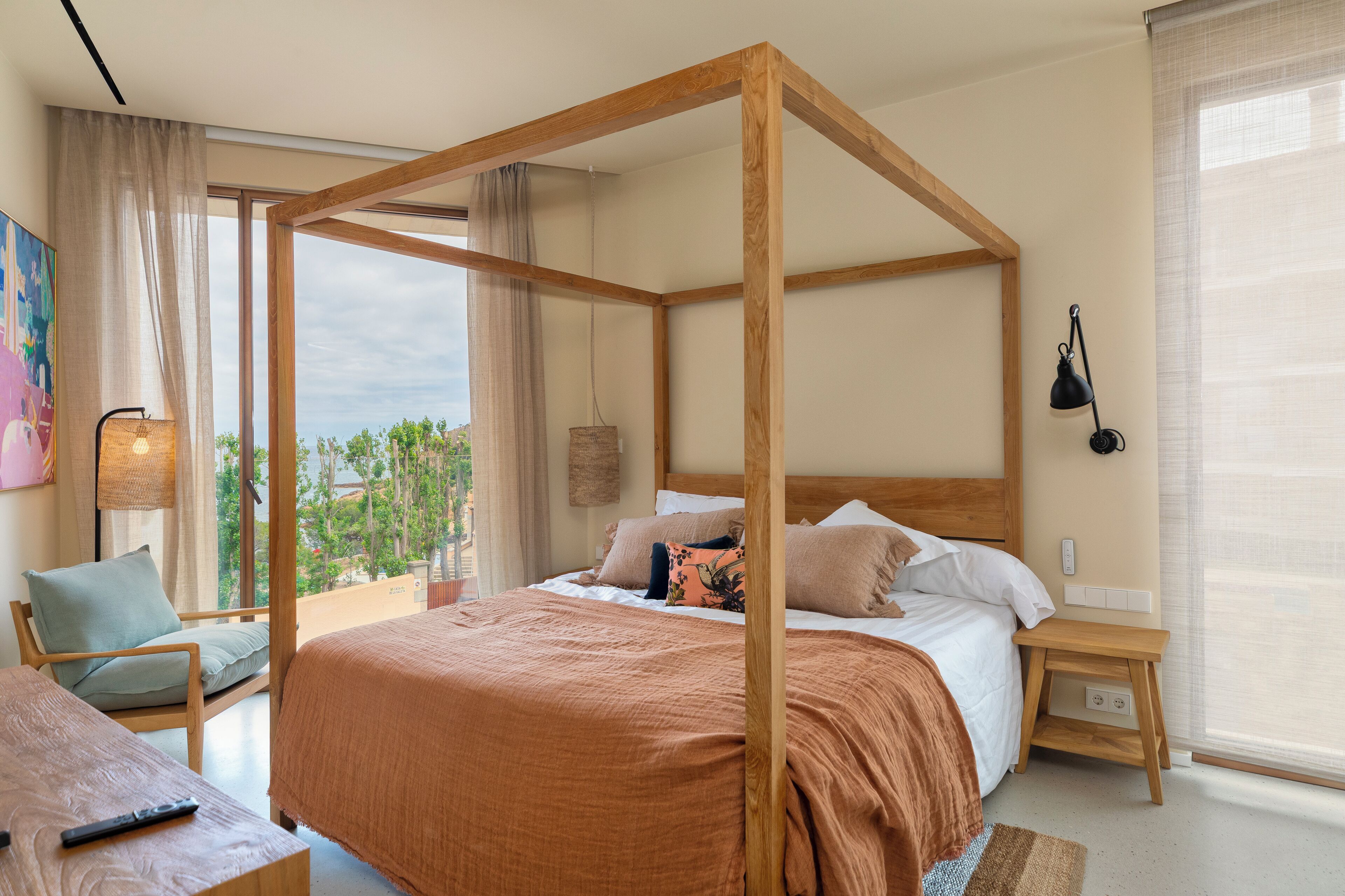 A bedroom with a four-poster bed, a wooden nightstand, a chair, and a view of greenery outside.