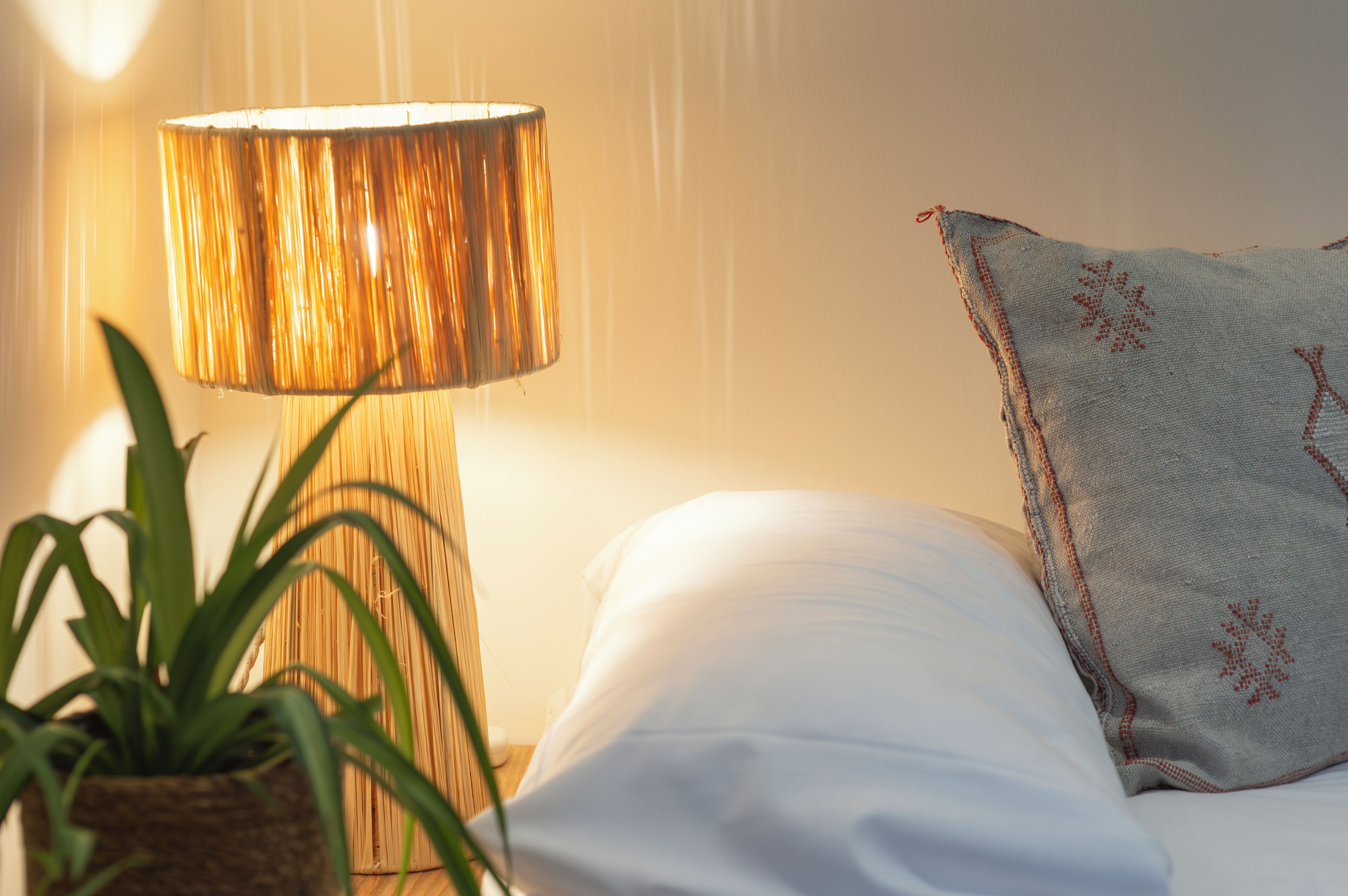 Un lit avec une literie blanche, une lampe avec un abat-jour texturé et un coussin décoratif orné de broderies.