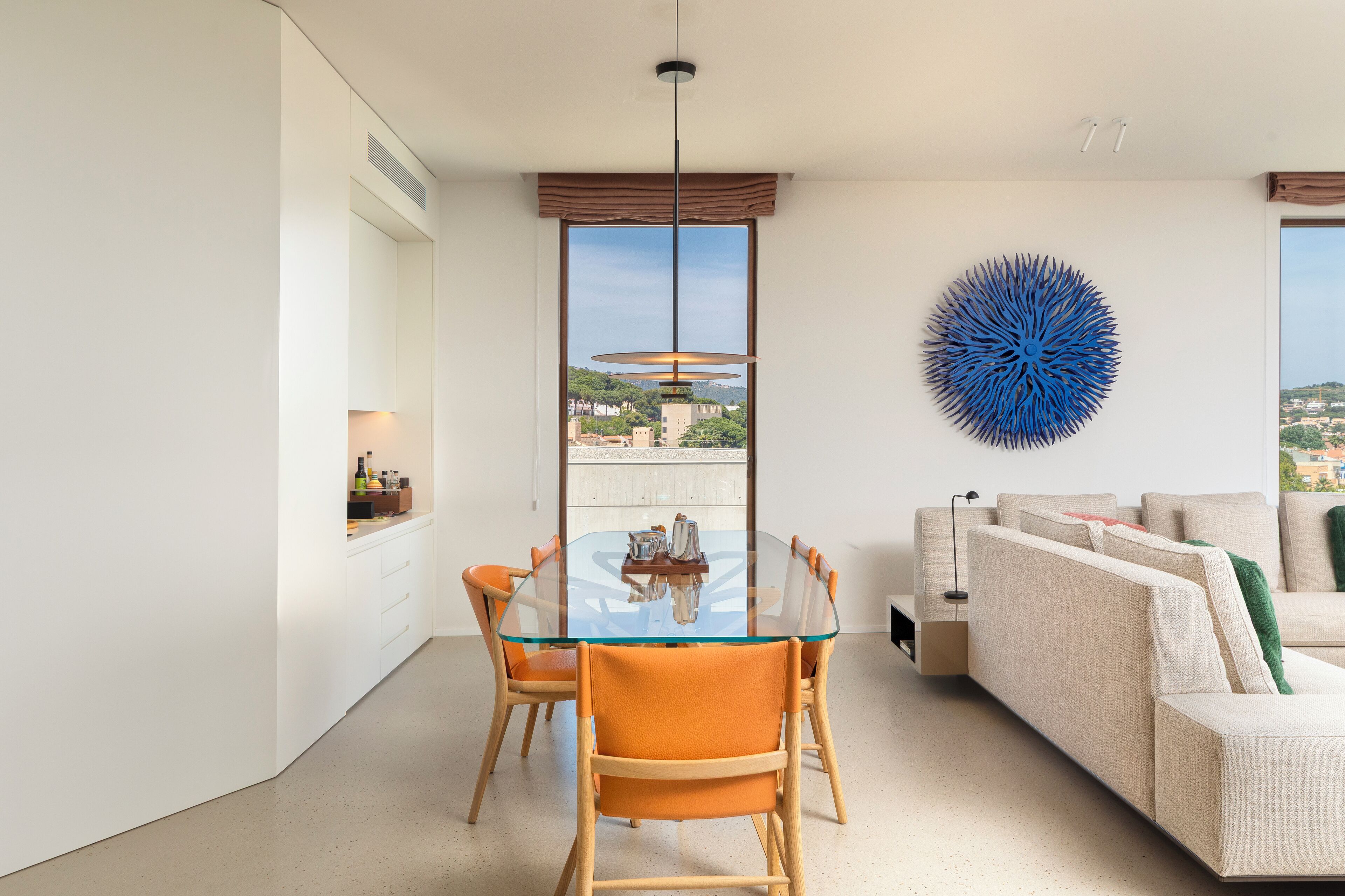 Ett modernt vardagsrum med ett glasbord, orange stolar, en beige soffa och en stor, blå väggdekoration.