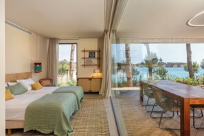 Quarto de hotel com duas camas, uma mesa de jantar com cadeiras, vista para o mar e uma varanda com mesa e cadeiras.