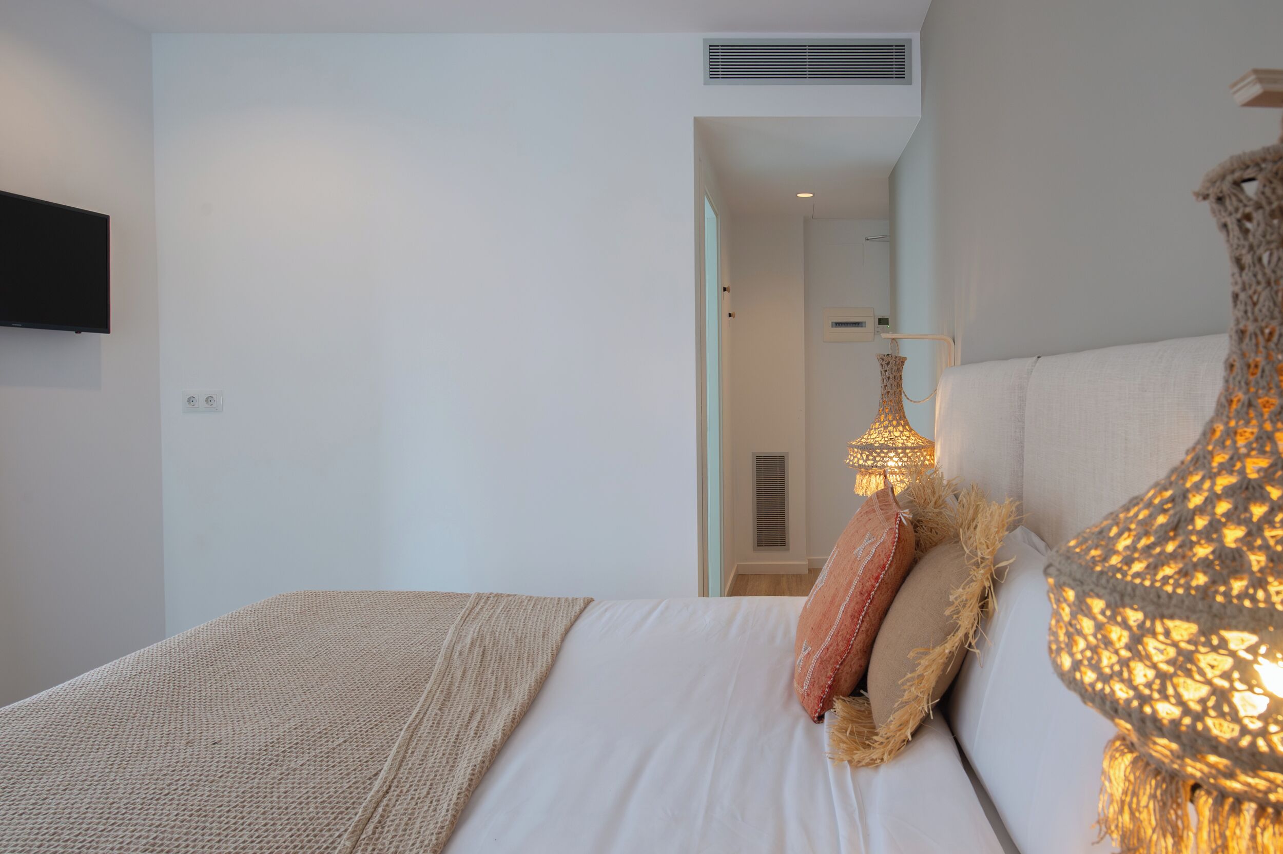 Un dormitorio con cama, televisión montada en la pared y lámpara con pantalla tejida.