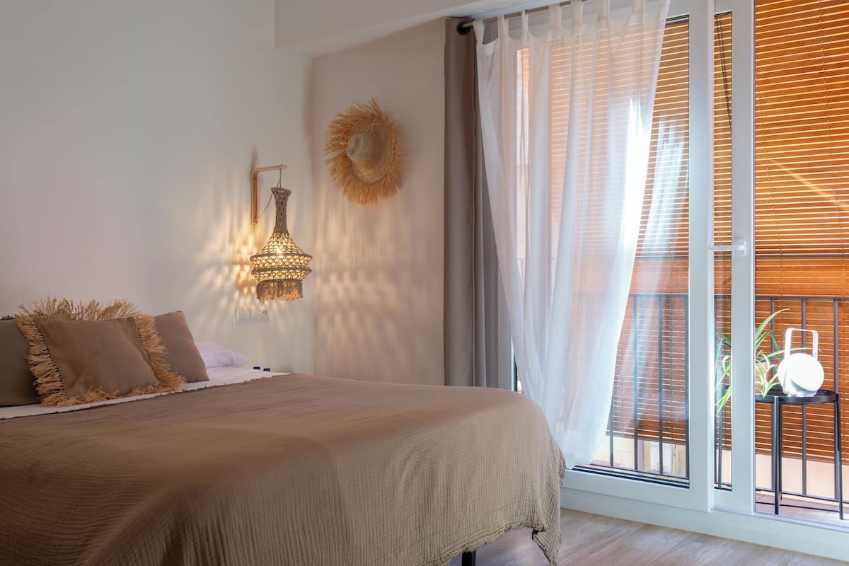 Een slaapkamer met een bed, een wandlamp, een decoratieve wandversiering en uitzicht door een raam met jaloezieën.