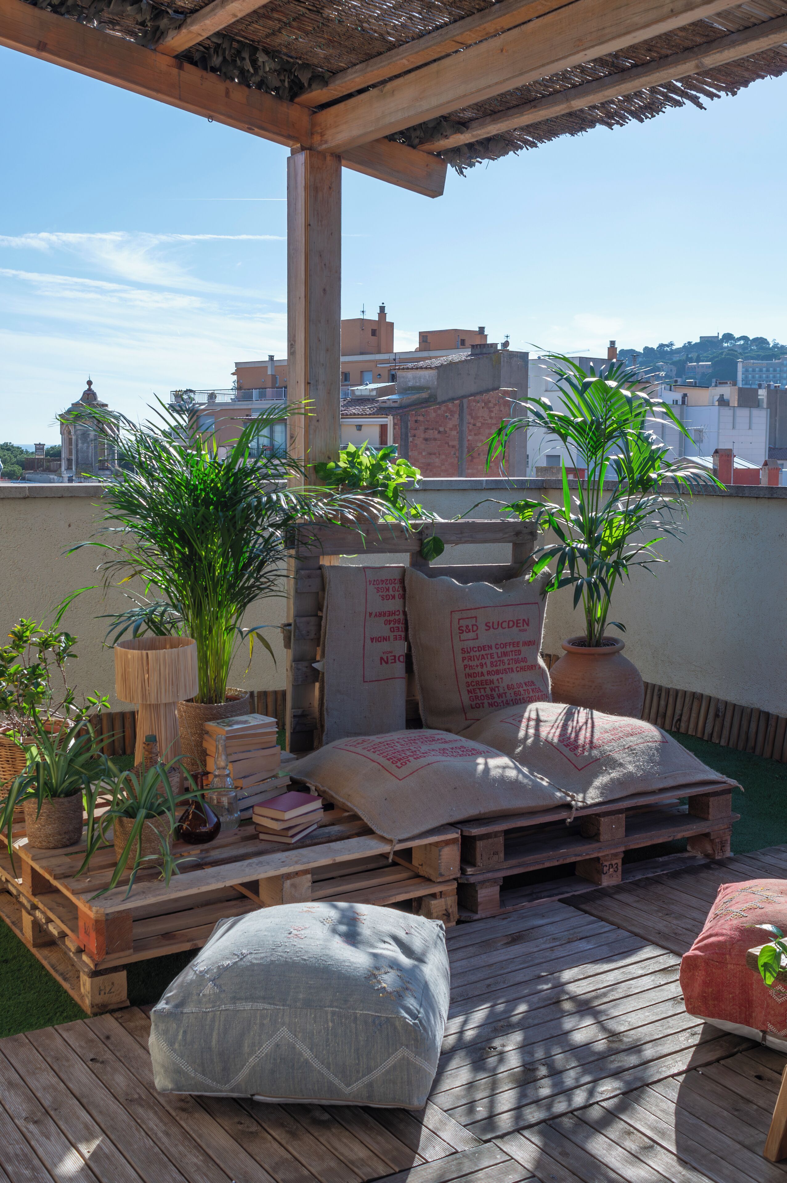 A rooftop terrace with wooden furniture, potted plants, and a view of buildings in the background.