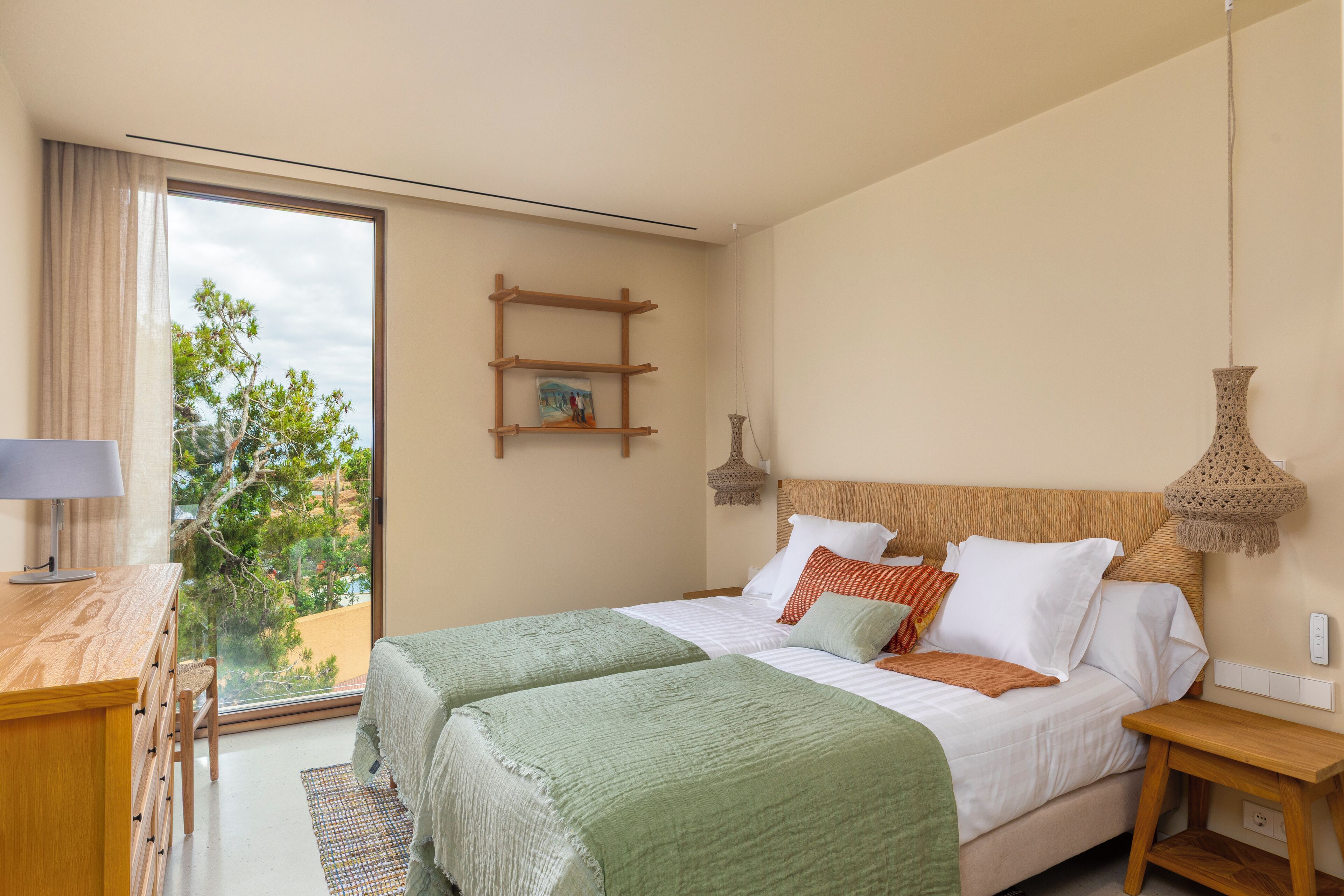 A bedroom with a large window, a wooden desk, a bed with green and white bedding, and a small shelf with a painting.