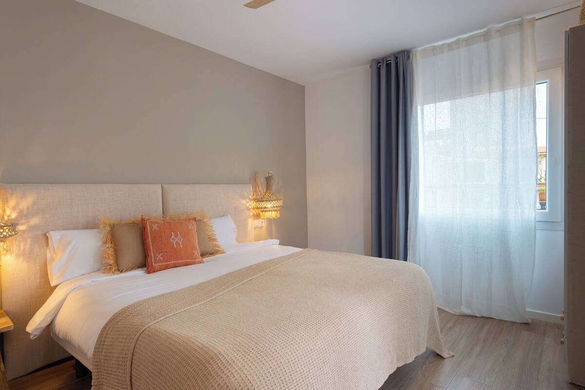 Een net opgemaakt bed met een beige deken en kussens, een bedlampje en een raam met dunne gordijnen.