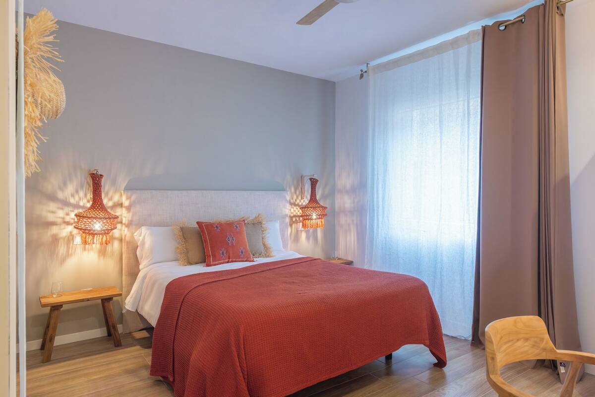 Een slaapkamer met een groot bed, twee bedlampen, een houten nachtkastje, een stoel en een raam met gordijnen.