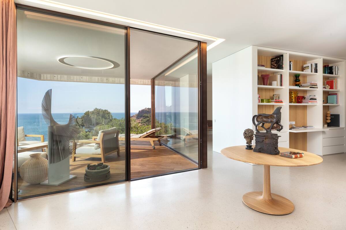 Een moderne woonkamer met een grote glazen deur die uitkomt op een balkon met uitzicht op zee. Er staat een houten tafel met decoratieve items en een boekenplank vol boeken en decoratieve objecten.