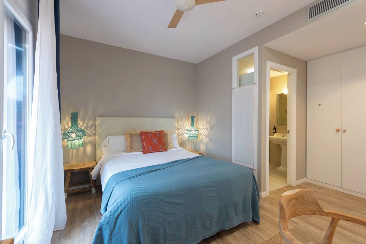 Een slaapkamer met een bed, nachtkastje, bankje, houten stoel, plafondventilator en een badkamer zichtbaar door een geopende deur.
