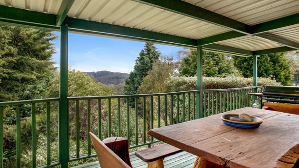 Una mesa y sillas de madera bajo un techo verde, con vista a un bosque y colinas.