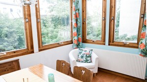 A room with a wooden table, a white chair with a floral pillow, and a view of greenery through large windows. - Marthas Veranda Apartment (Vienna)