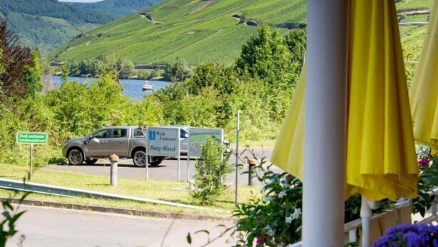 A view of a hotel with a mountain backdrop, a parked truck, and yellow umbrellas. - Hotel Weinhaus Moselblick (Burg)