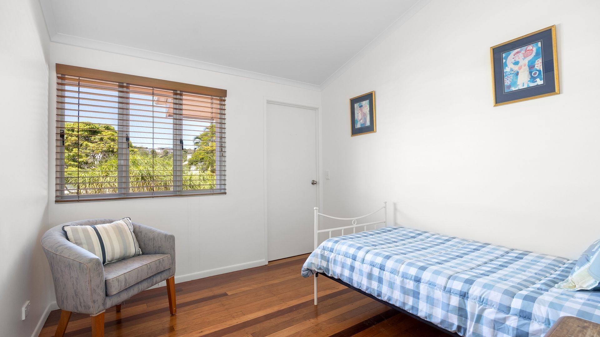 Un dormitorio con una cama, una silla, un ventanal con persianas y dos cuadros enmarcados en la pared.