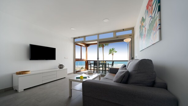 A modern living room with a large window, a flat-screen TV, and a grey sofa. - Apartamentos Los Papagayos (San Bartolomé de Tirajana)