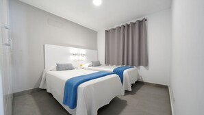 A hotel room with two beds, a large window with curtains, a bedside table with a lamp, and a wall-mounted light fixture. - Apartamentos Los Papagayos (San Bartolomé de Tirajana)
