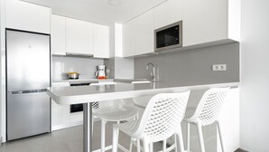 A modern kitchen with white cabinetry, a stainless steel refrigerator, a microwave, and a bar area with white chairs. - Apartamentos Los Papagayos (San Bartolomé de Tirajana)