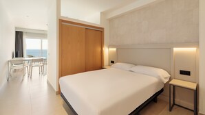 Habitación de hotel moderna con una cama grande, mesitas de noche, un escritorio con sillas y vistas al mar a través de un ventanal amplio.