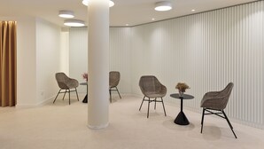 Un moderno vestíbulo de hotel con techo alto, iluminación empotrada y una columna grande. Hay cuatro sillas de mimbre alrededor de una pequeña mesa redonda con un florero.