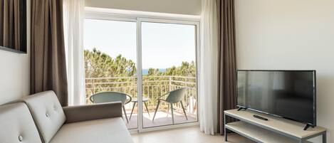 A modern hotel room with a sofa, a TV on a stand, and a balcony with chairs and a view of trees.
