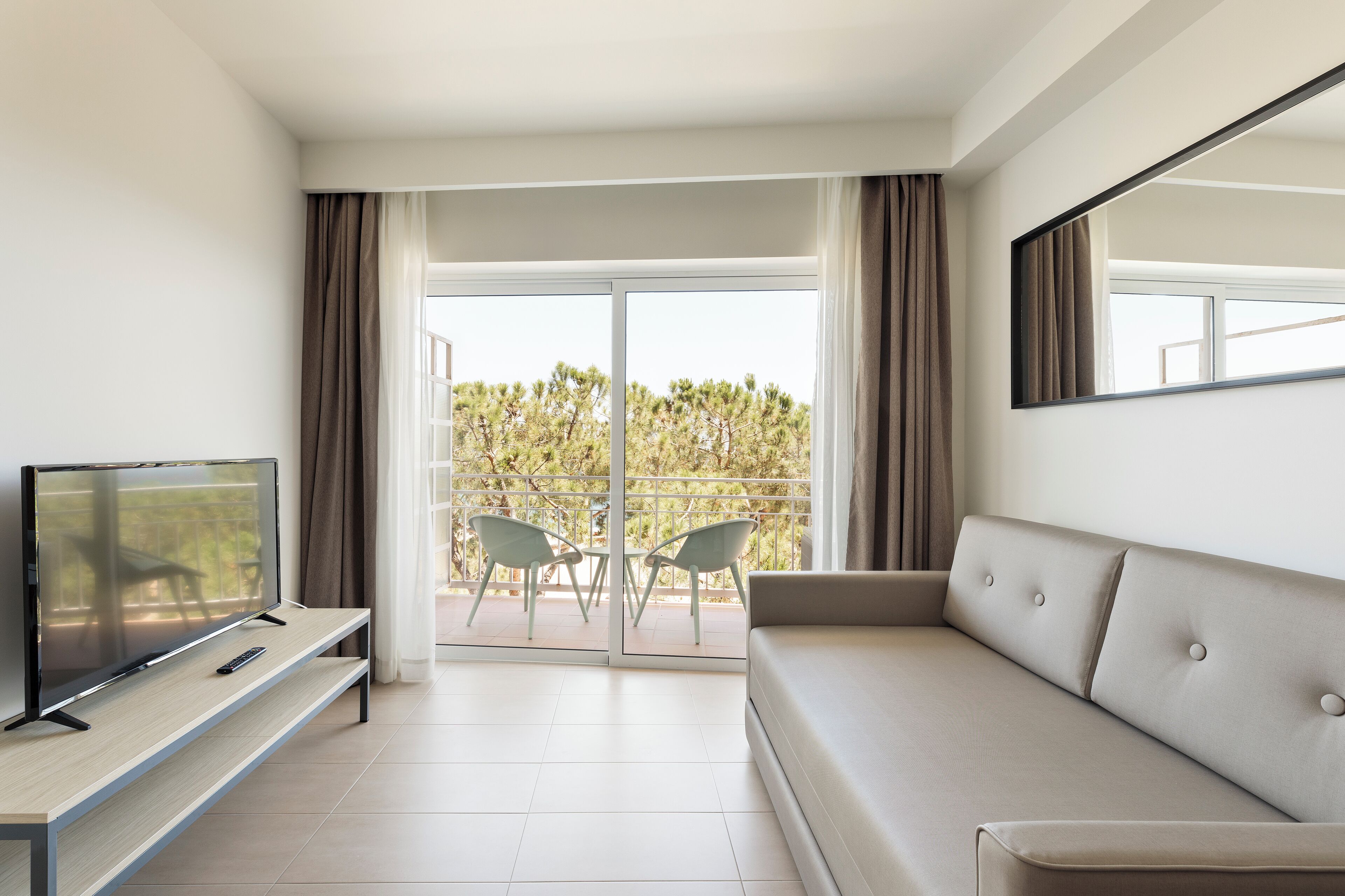 Habitación de hotel moderna con televisor de pantalla plana sobre un mueble de madera, un sofá color beige y un balcón con dos sillas.