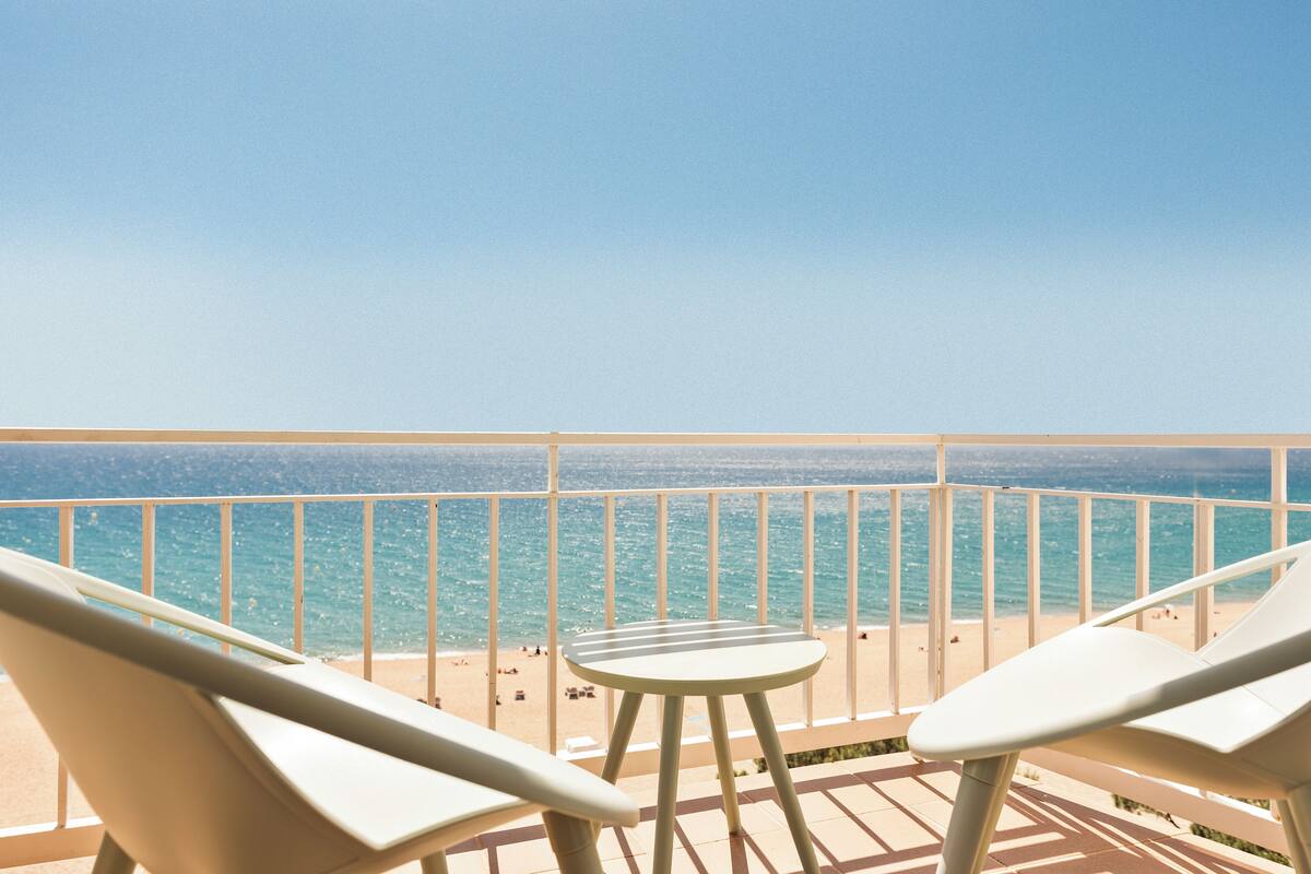 Un balcon avec deux chaises blanches et une petite table, donnant sur une plage où se trouvent des gens et sur l’océan.