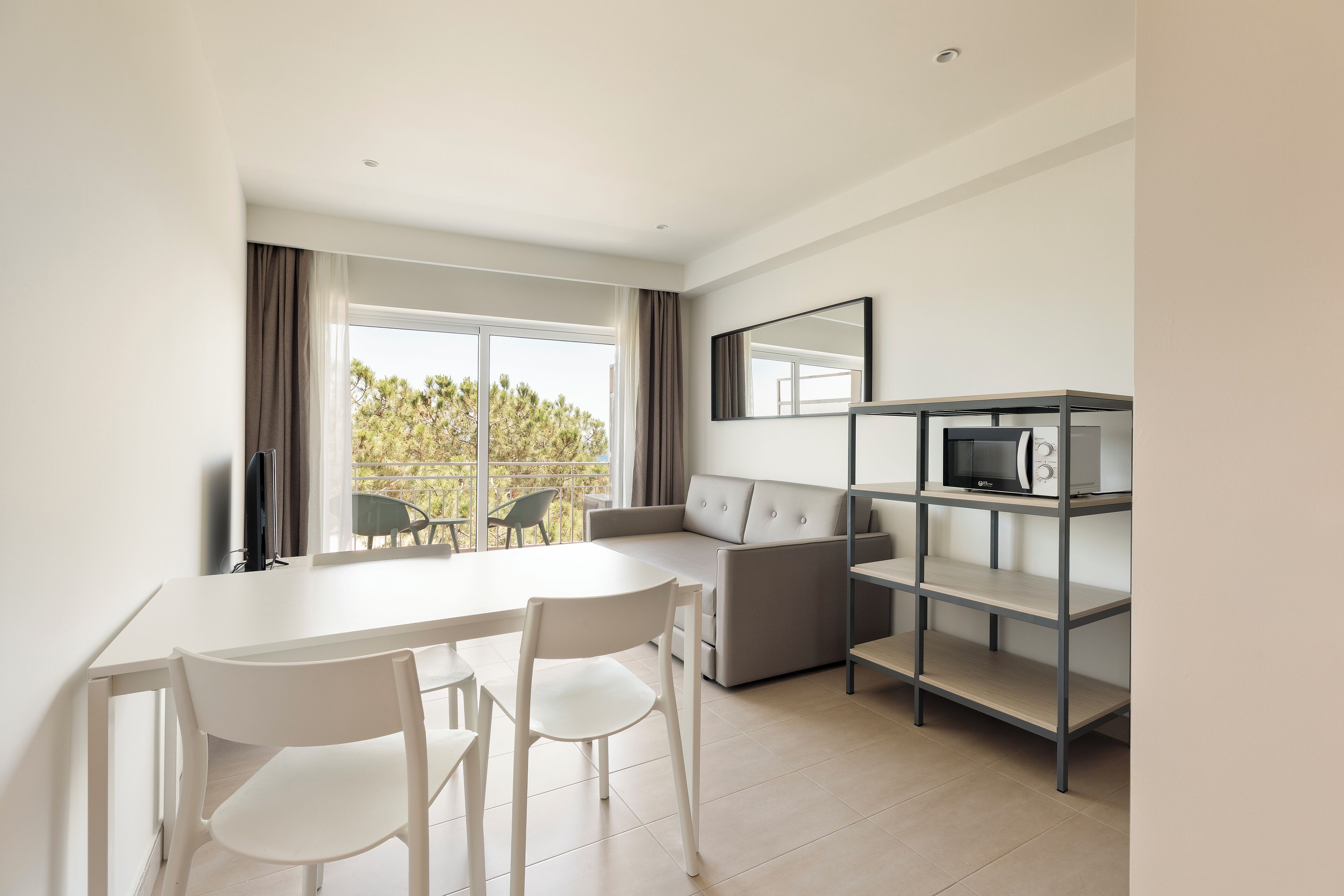 A modern living room with a white dining table, two white chairs, a grey sofa, a TV on a shelf, and a large window with curtains.