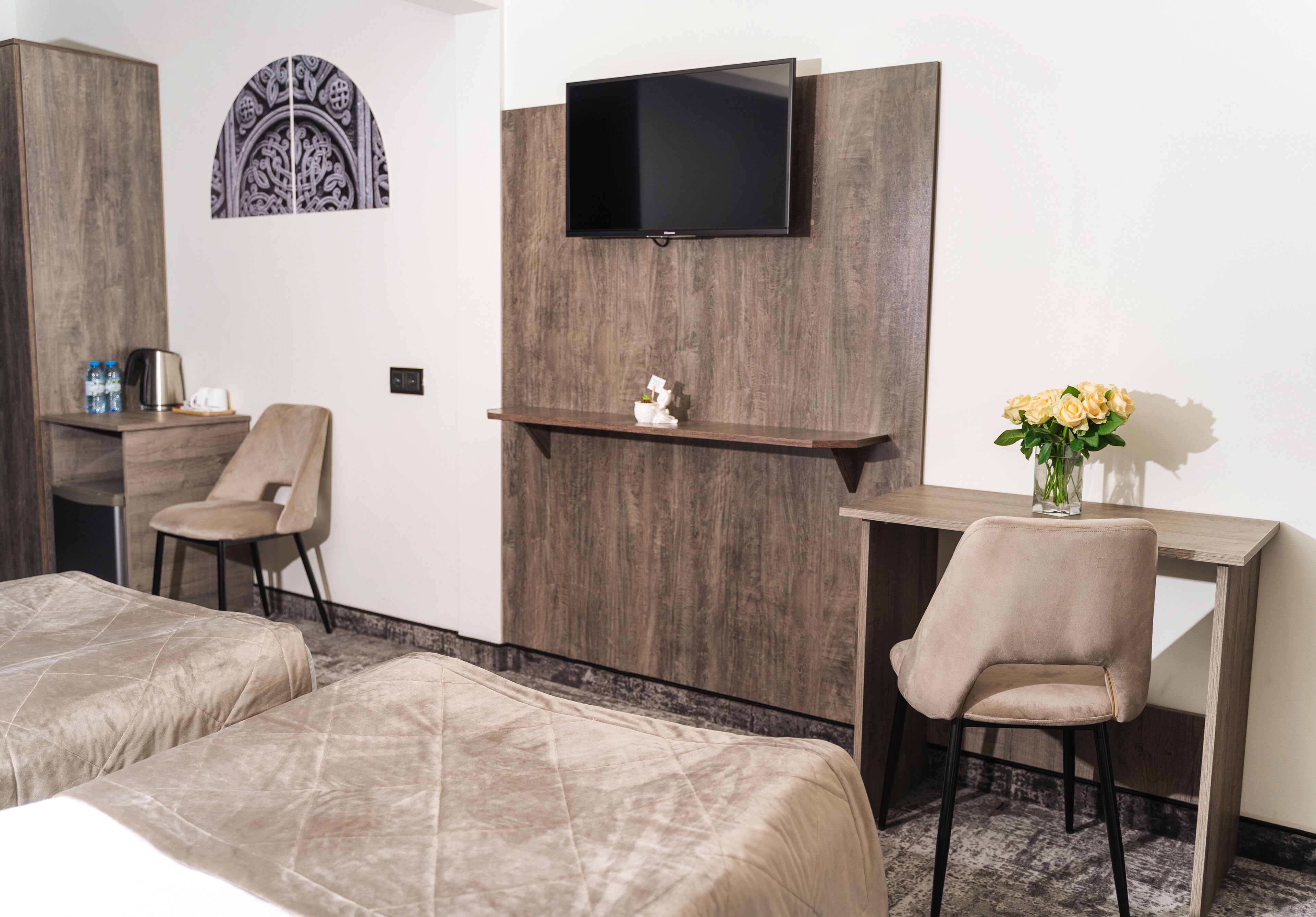 A hotel room with two beds, a desk, a chair, a TV mounted on a wooden panel, and a small shelf with a vase of flowers.