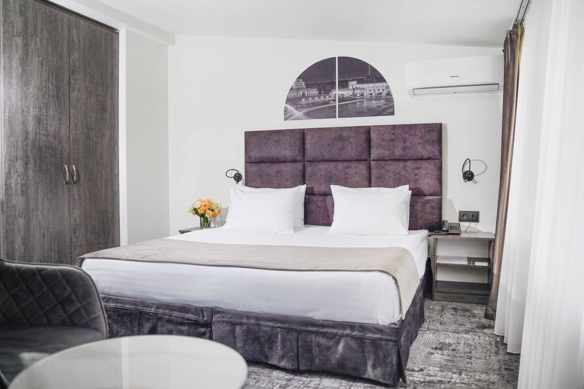 A hotel room with a large bed, a wardrobe, a bedside table with a lamp, and a view of a cityscape.