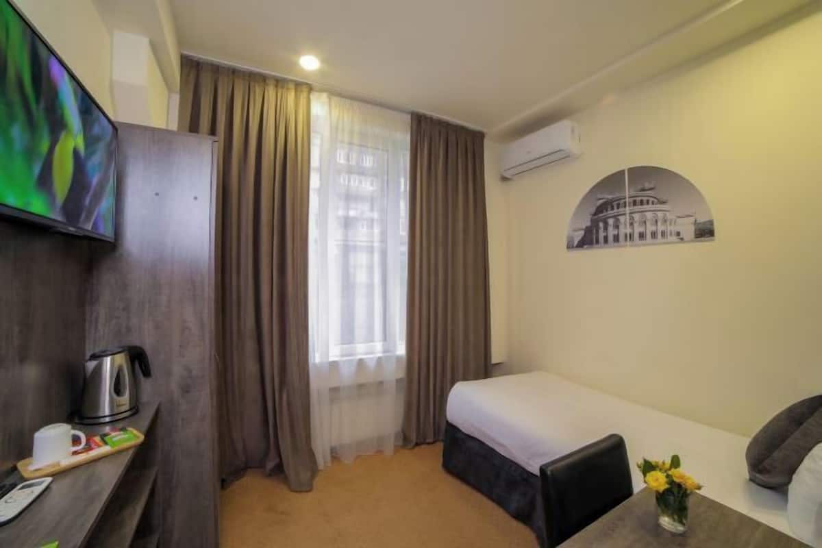 A hotel room with a bed, a desk with a kettle, a TV, and a wall-mounted air conditioner.
