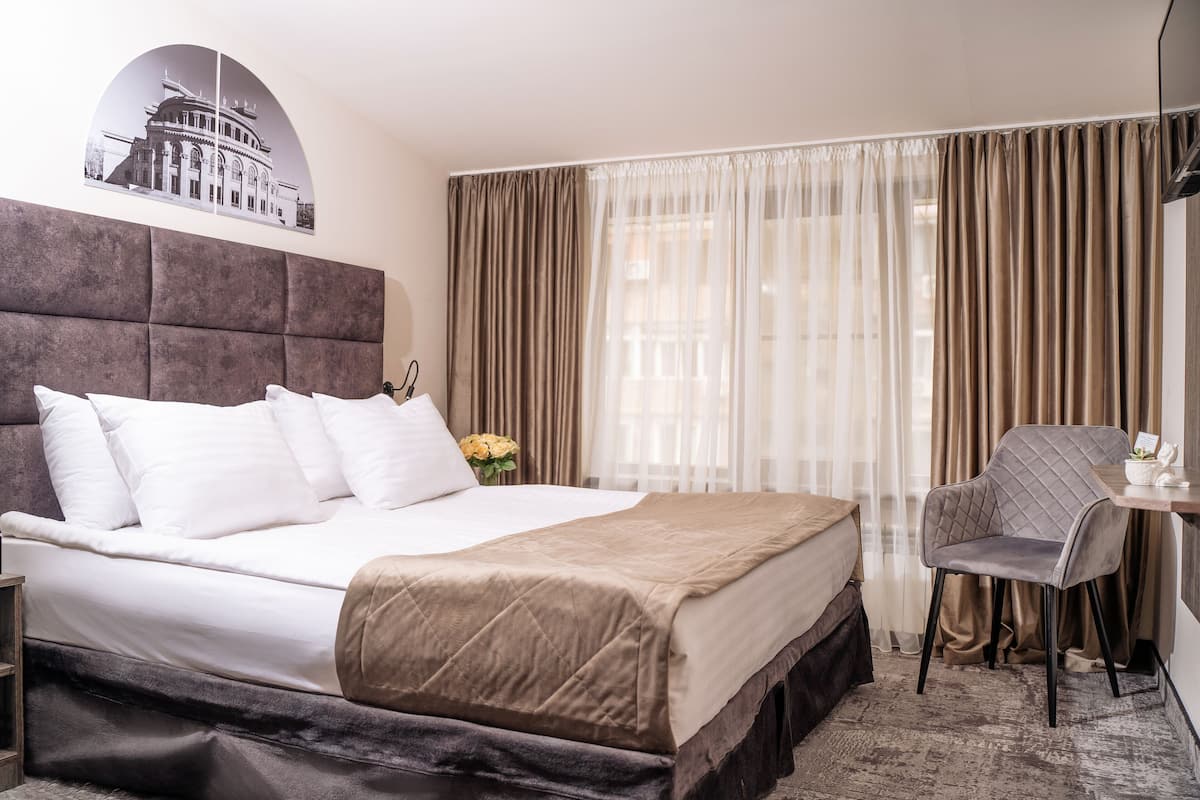 A hotel room with a large bed, a headboard, a desk with a chair, and a window with curtains.