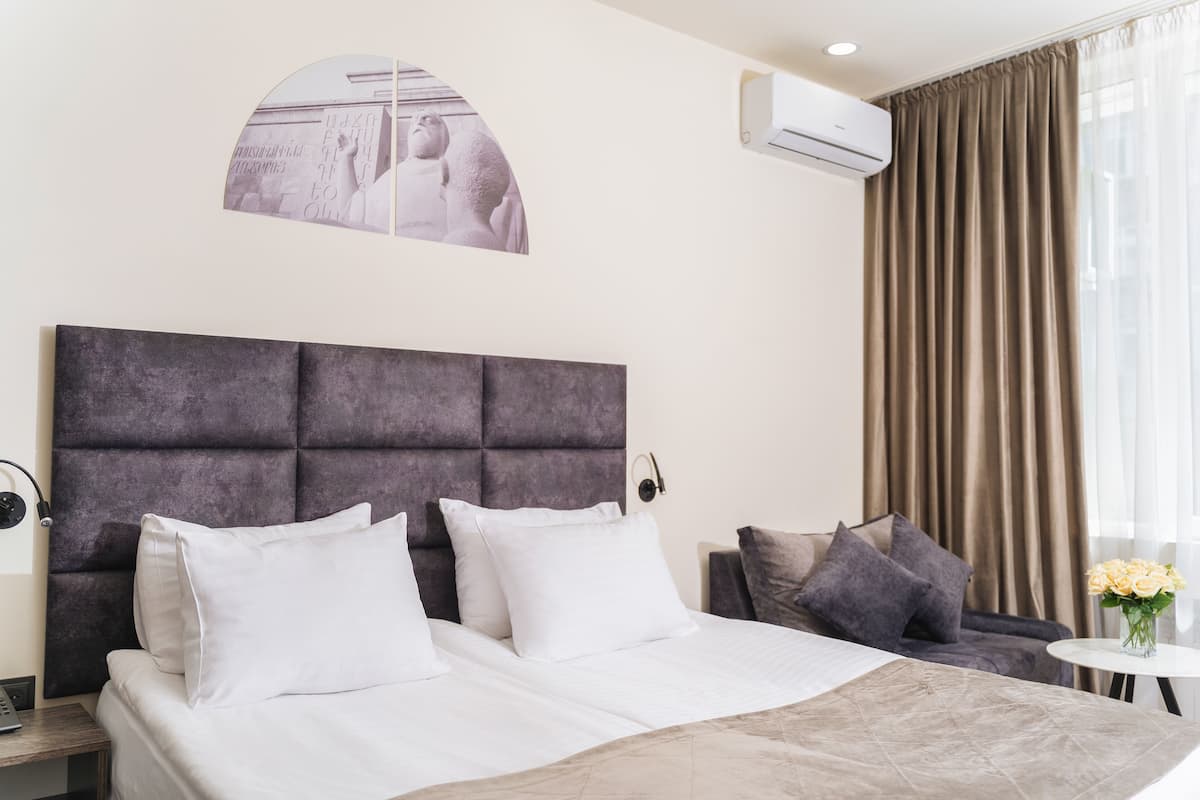 A hotel room with a large bed, a headboard, a small table with a vase of flowers, a sofa, and a wall-mounted air conditioner.