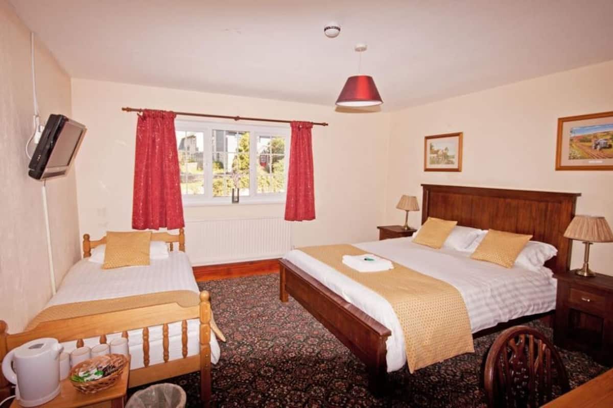 Quarto duplo com duas camas, televisão, janela com cortinas vermelhas, cabeceira de madeira e uma luminária.