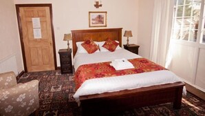 Un dormitorio ordenado con una cama de madera, dos mesitas de noche con lámparas, un tapete estampado, un sillón floral y una puerta con un aviso.