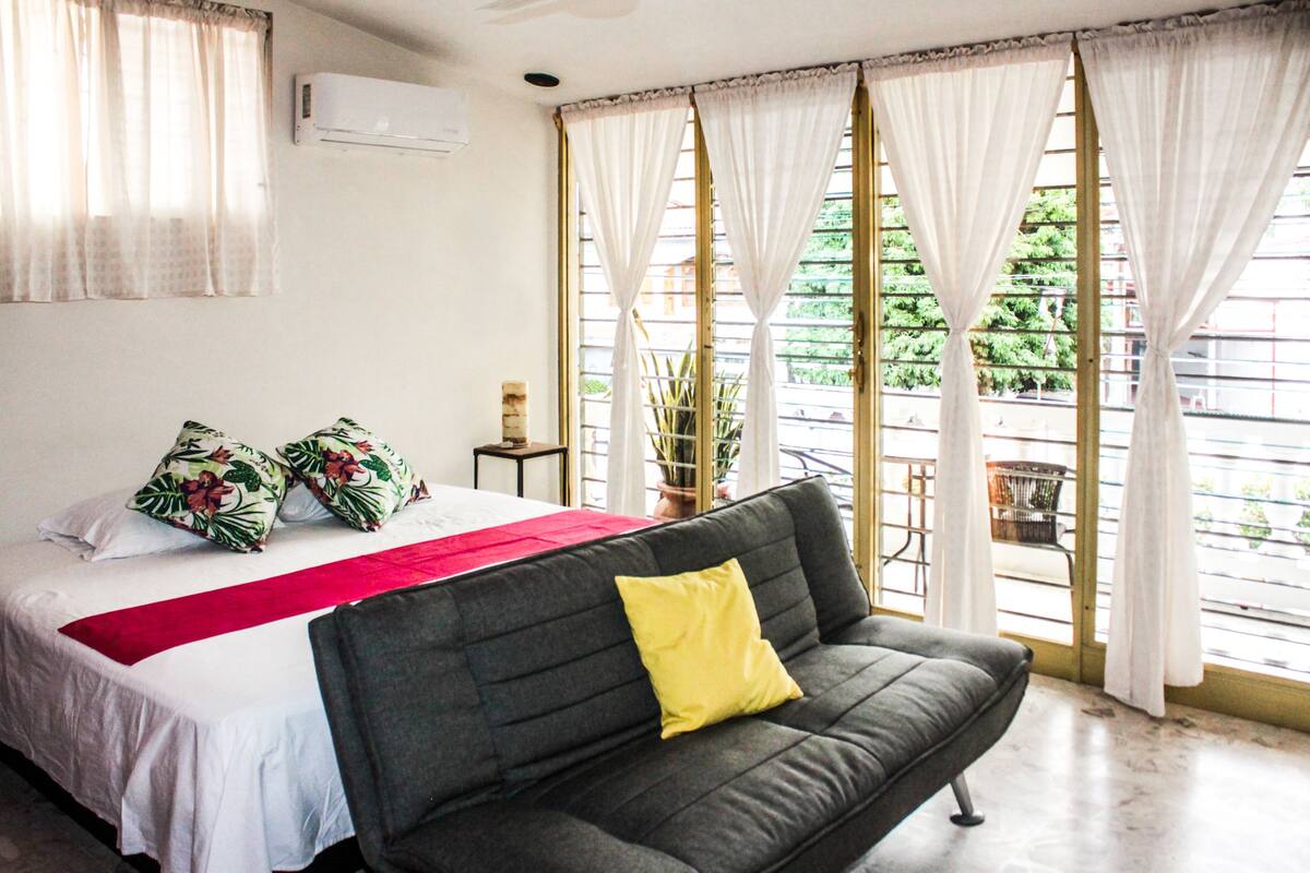 Ein Schlafzimmer mit einem Bett, einem grauen Sofa mit gelbem Kissen, einer roten Decke und einem Fenster mit Vorhängen.