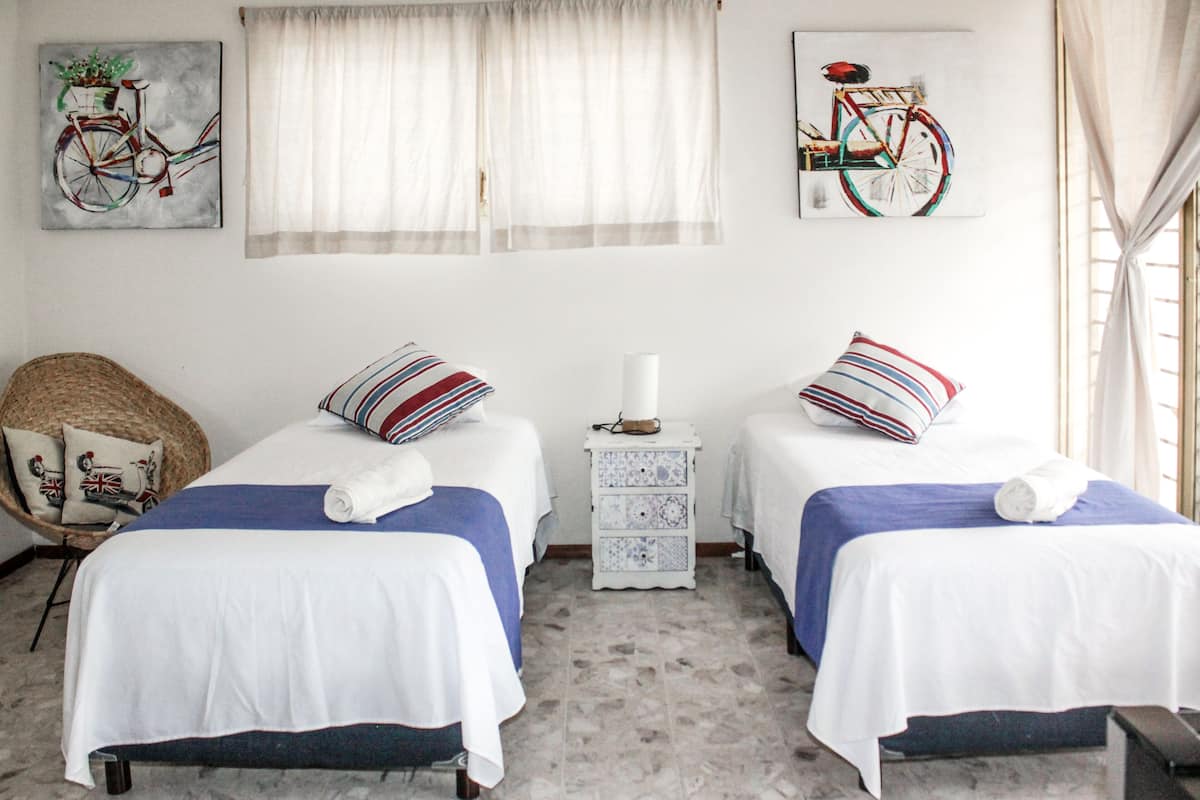 Ein Zimmer mit zwei Betten, einem Korbsessel, einem kleinen weißen Nachttisch und Bildern an den Wänden.
