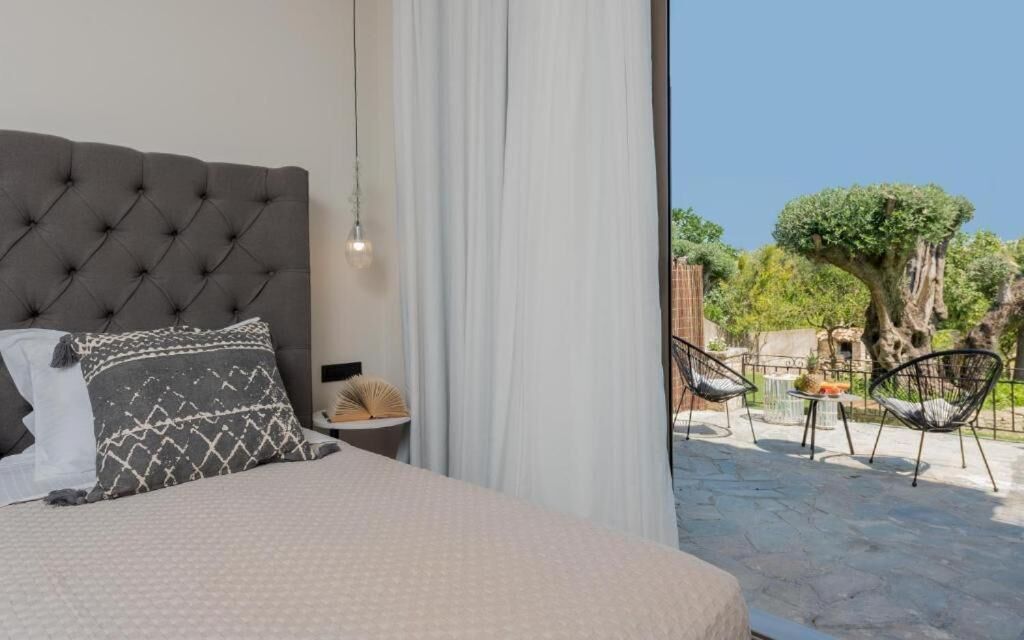A bedroom with a tufted headboard, a bed with patterned pillows, a bedside table with a lamp, and a view of an outdoor patio with chairs and a tree.