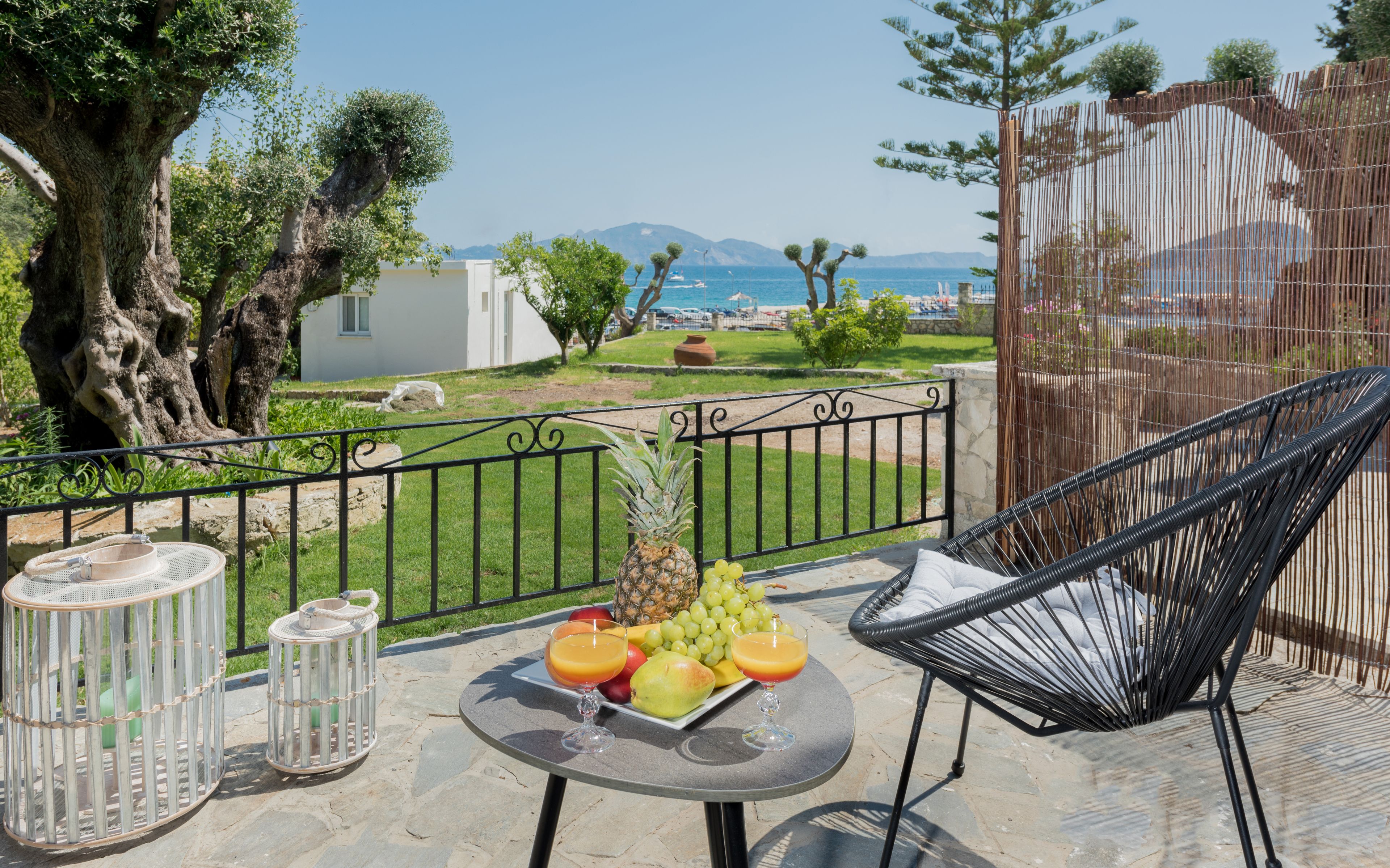 A balcony with a table set with fruit and drinks, a wicker chair, and a view of the sea and mountains.