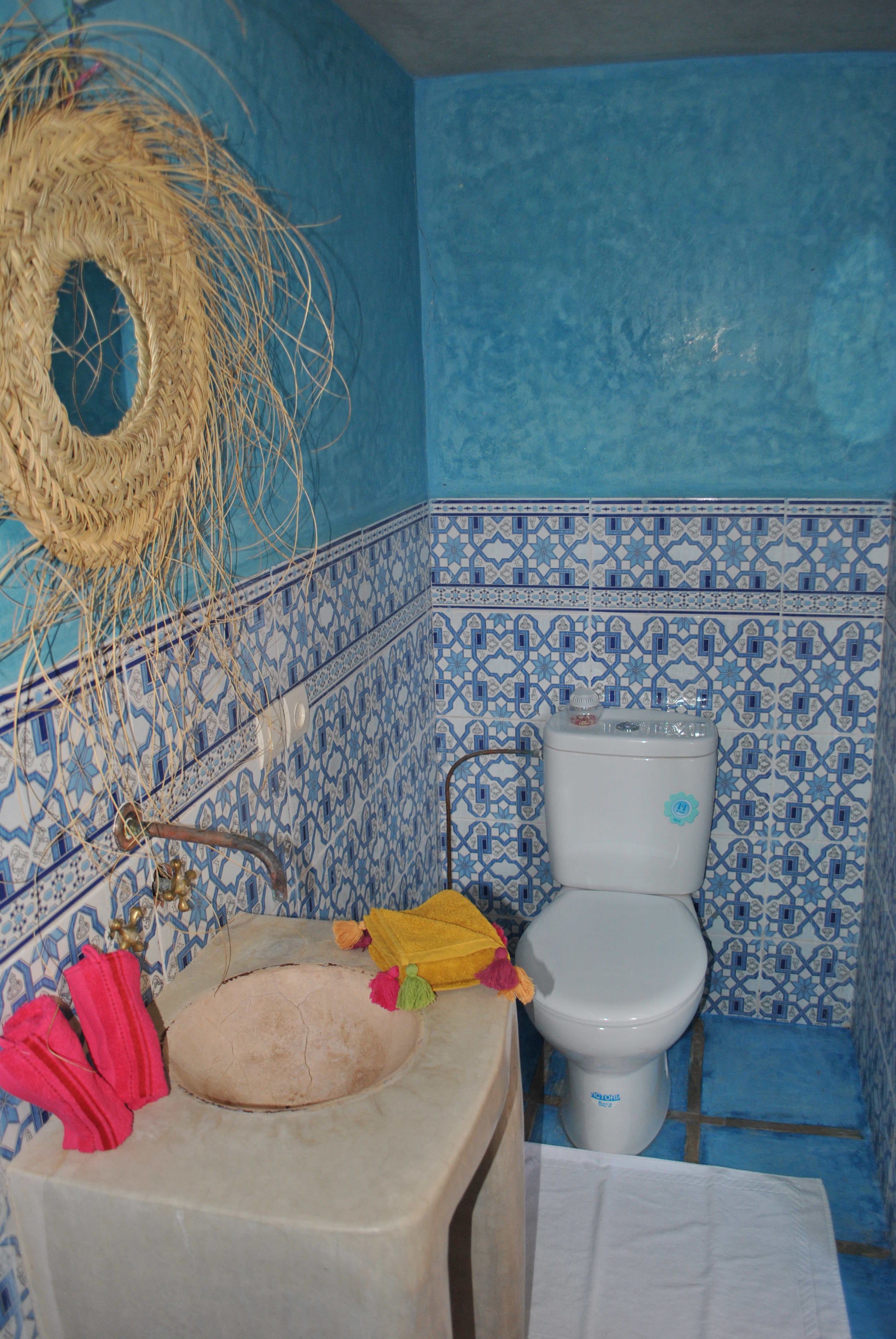 A bathroom with a blue tiled wall, a white toilet, a sink with a yellow towel, and a straw basket hanging on the wall.