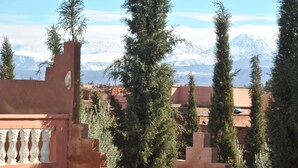 A view of a mountainous landscape with snow-capped peaks in the background, a building with a reddish-brown facade, and tall evergreen trees in the foreground. - Auberge Kasbah Darla (Aghouatim)