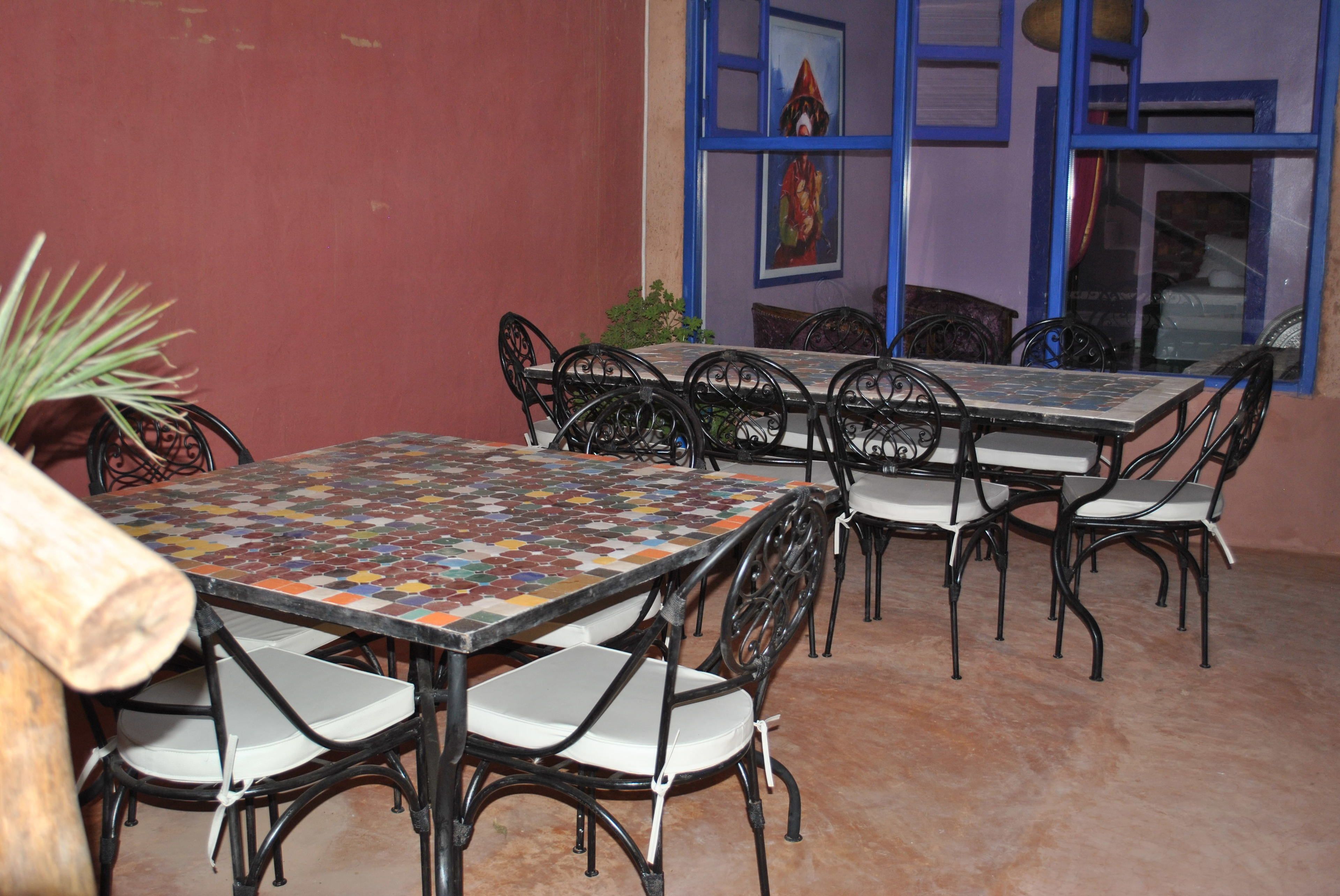 Un patio con una mesa y sillas de cerámica, una planta en maceta y una ventana con marco azul que muestra un cuadro.