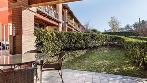 Un espace patio avec une table en verre et des chaises en osier, un bâtiment en briques doté d’un balcon et une haie bien entretenue.
