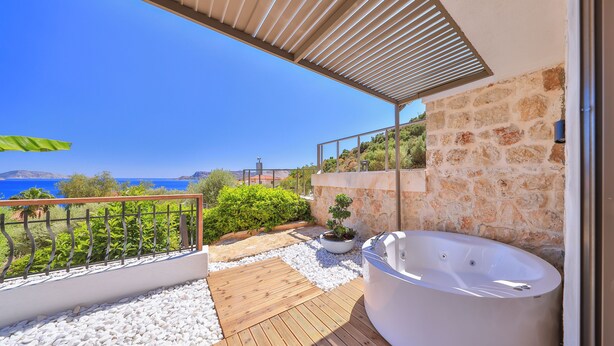 A modern outdoor deck with a stone wall, a wooden deck, a white hot tub, and a view of the sea. - Molino Hotel (Kas)
