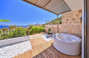 A modern outdoor deck with a stone wall, a wooden deck, a white hot tub, and a view of the sea. - Molino Hotel (Kas)