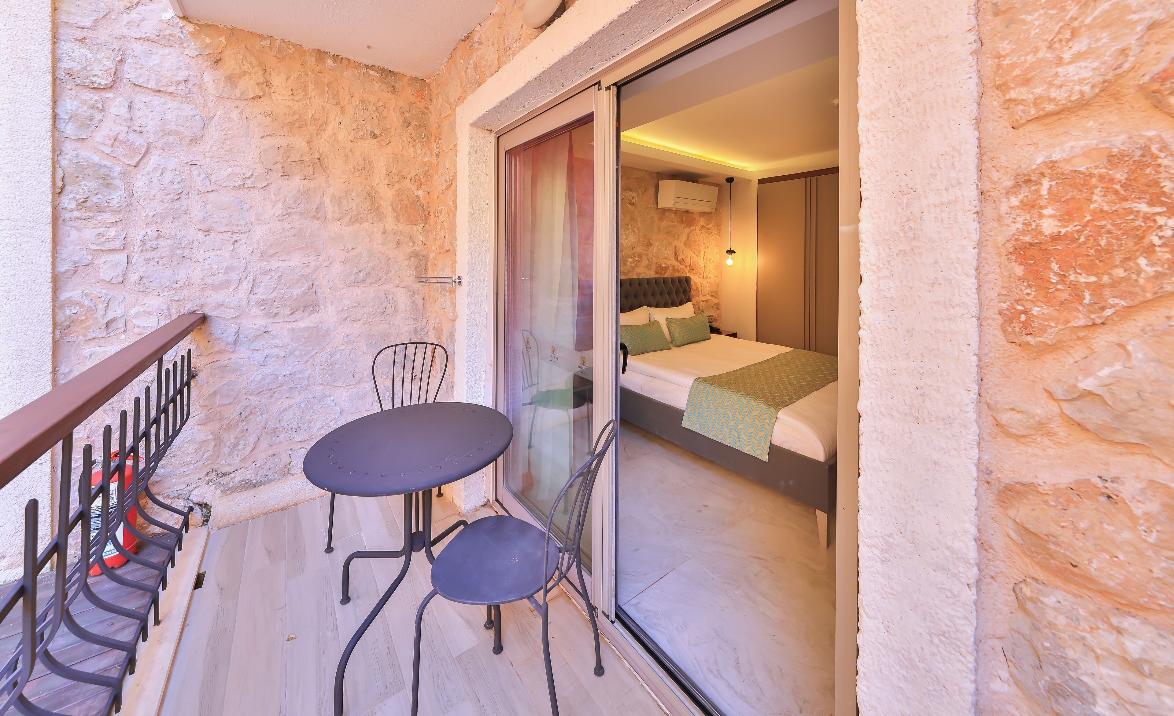 A balcony with a table and chairs, a sliding glass door leading to a bedroom with a bed and a nightstand.