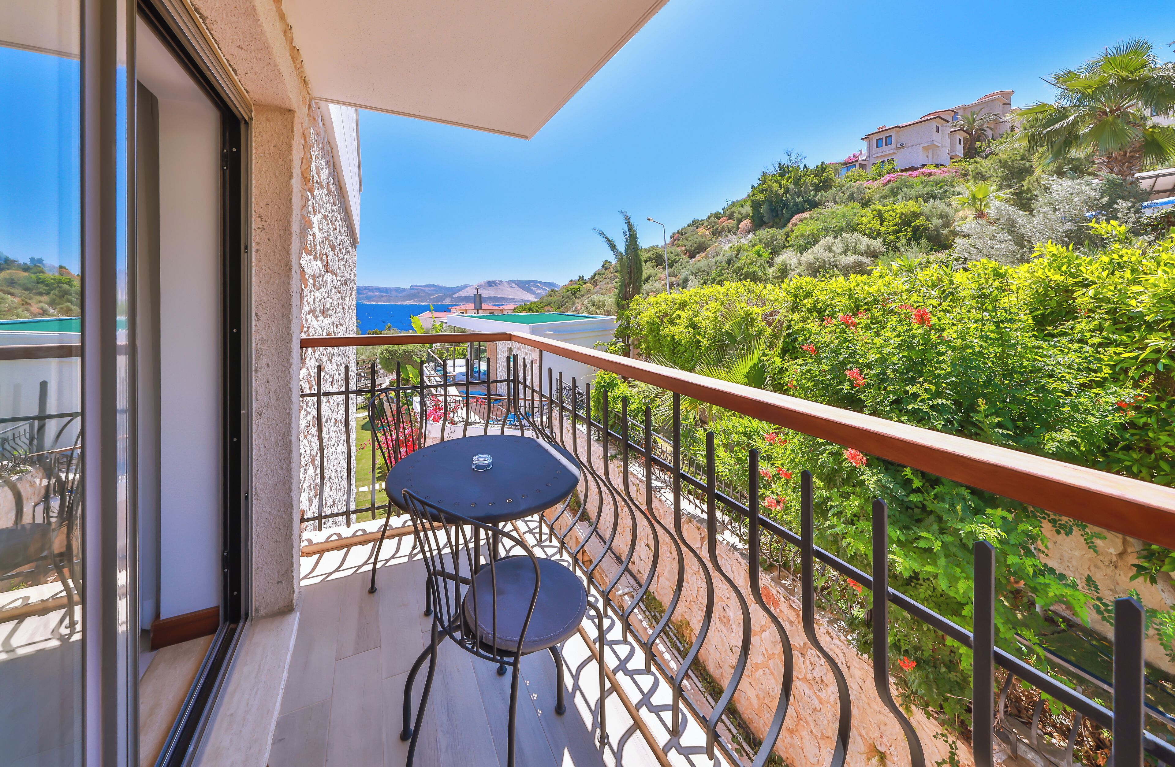 A balcony with a table and chairs, overlooking a coastal landscape with vegetation and a building in the distance.