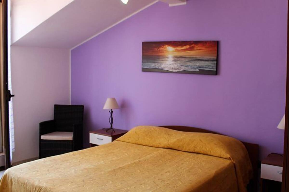Une chambre avec un lit, une chaise, une table de chevet et un tableau représentant un coucher de soleil sur l’océan.