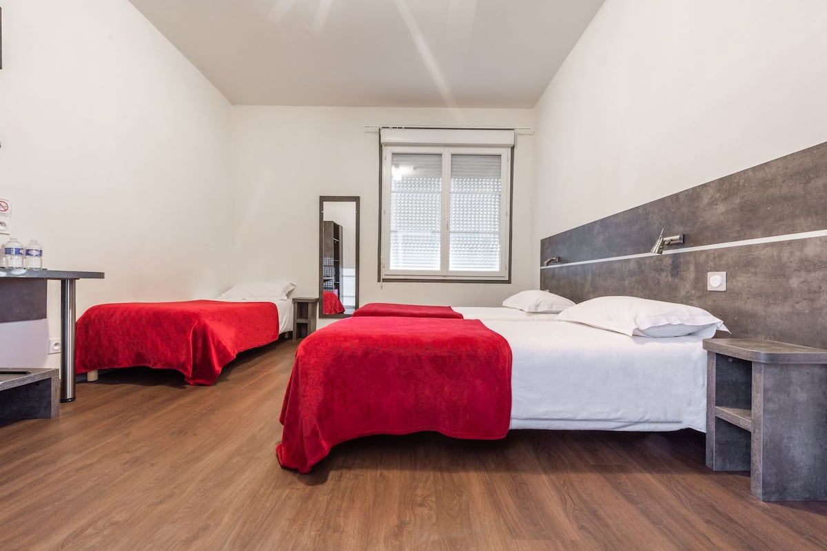 A hotel room with two beds, a large window with blinds, a mirror, and a wooden floor.