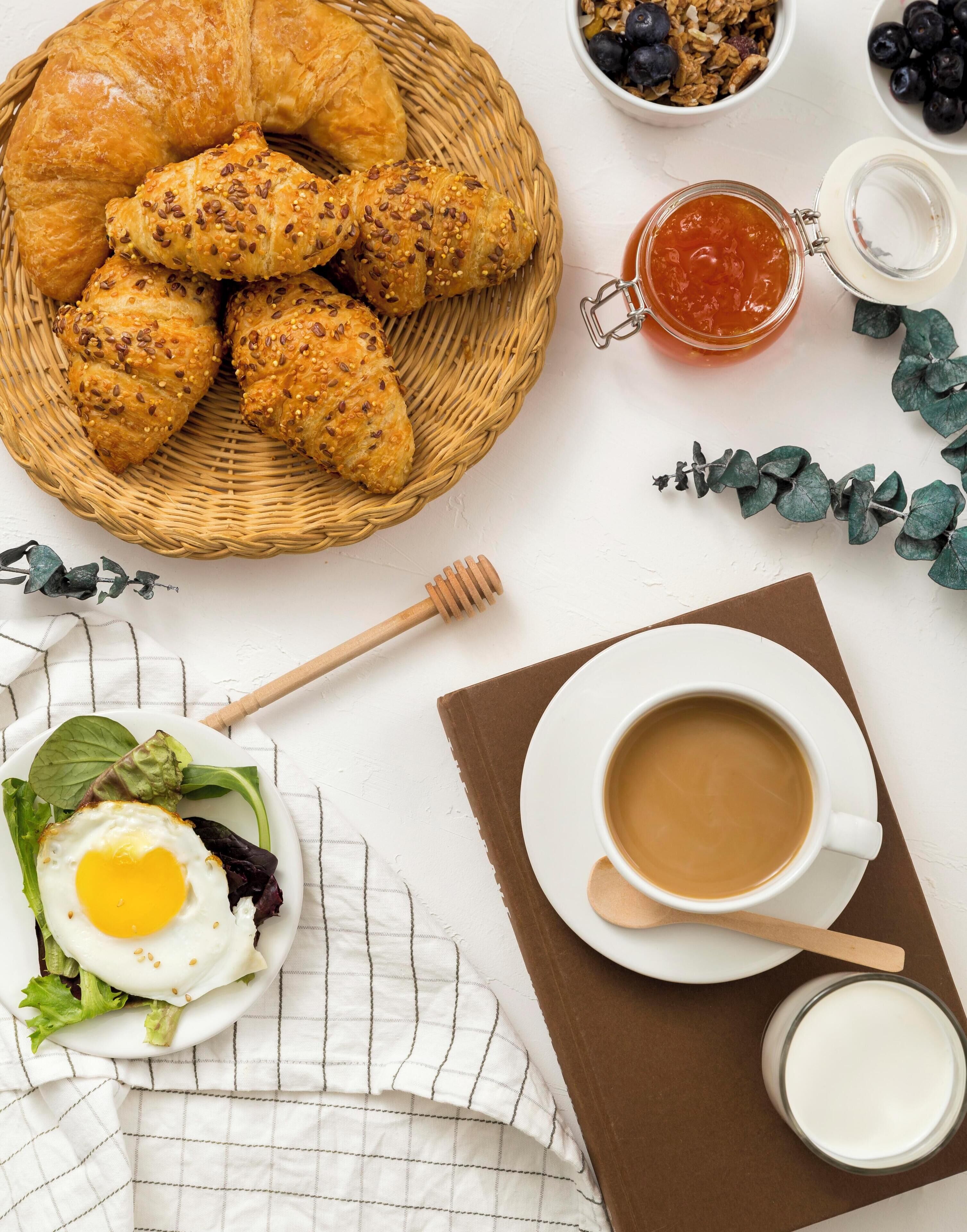 Een ontbijttafel met croissants, een kom granola met bosbessen, een potje jam, een kop koffie, een bord met een ei en groenten, en een honinglepel.