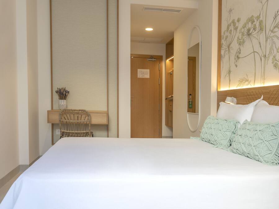 A hotel room with a large bed, a wooden headboard, a wall-mounted mirror, a small desk with a chair, and a wall art piece.