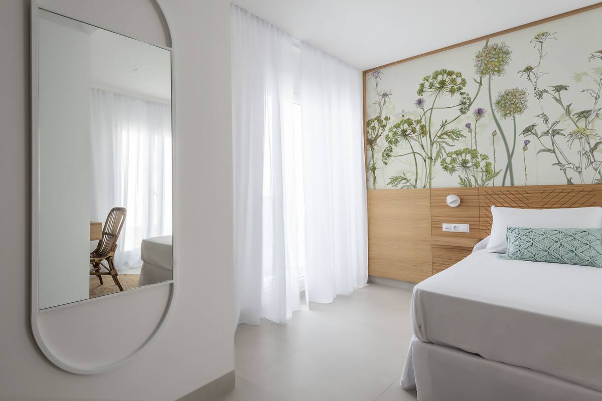 Une chambre d’hôtel avec un grand miroir, un lit avec une literie blanche, une tête de lit en bois et un papier peint à motifs floraux.