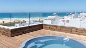 Zwembad op een dak met een houten terras en een glazen balustrade, met uitzicht op een strand en stadsbeeld.